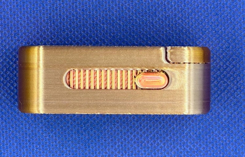 May include: A rectangular, metallic gold-toned object featuring a horizontal recessed channel. Inside the channel is a textured, ribbed copper-coloured sliding mechanism with a small, oval-shaped button positioned on the right side.
