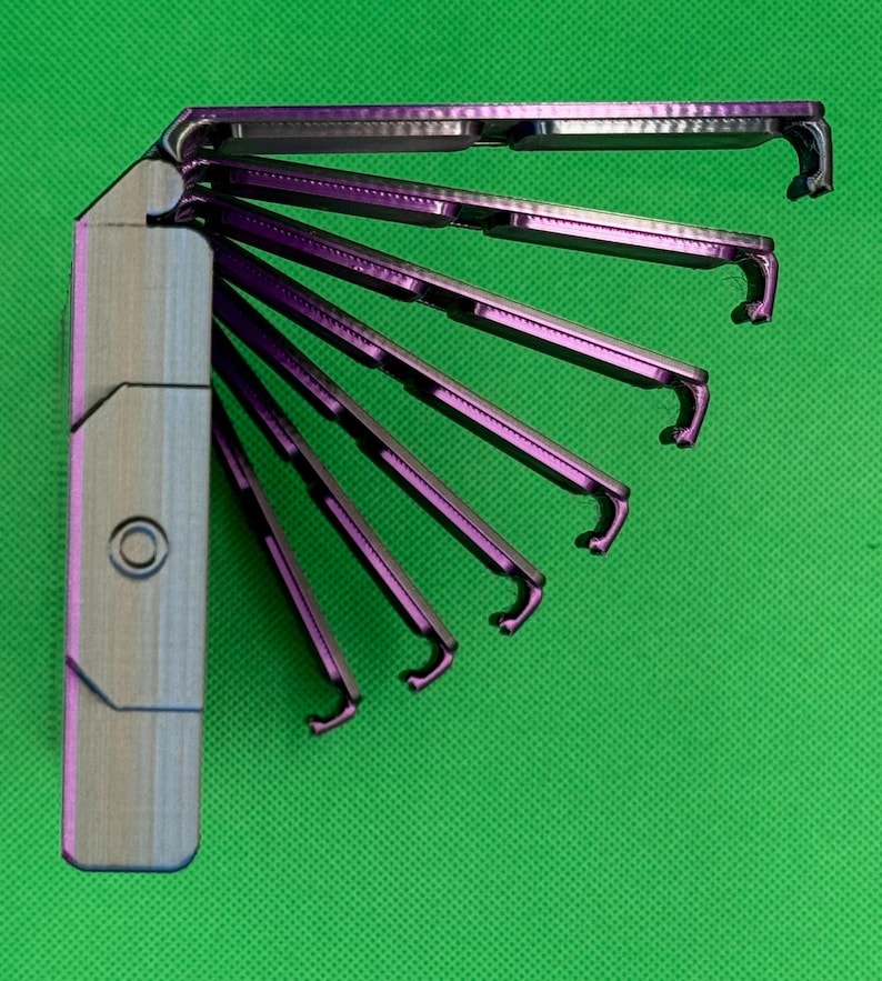 May include: A metallic purple multi-tool with a fan-like design, featuring six individual folding arms that extend from a central rectangular handle. Each arm has a hooked end, and the handle displays a circular indentation on its side.