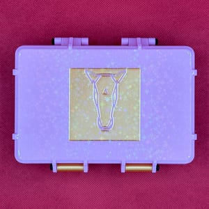 May include: A rectangular, lavender-colored case with a gold-colored horse head design in the center. The case has a textured surface with star-shaped accents and gold-colored clasps.