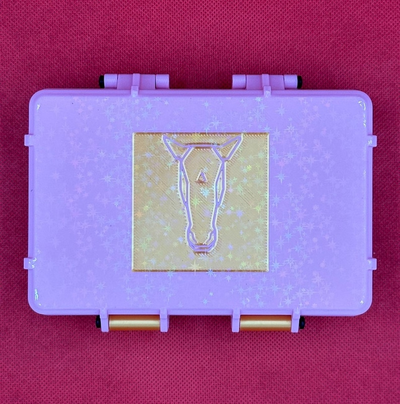 May include: A rectangular, lavender-colored case with a gold-colored horse head design in the center. The case has a textured surface with star-shaped accents and gold-colored clasps.