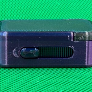 May include: A small, rectangular, dark purple device with a sliding control and a black top. The device is set against a green background.
