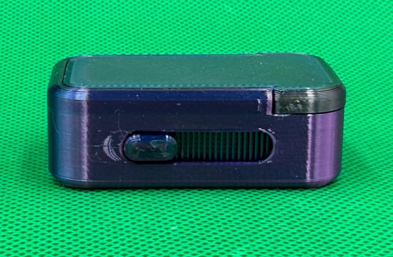 May include: A small, rectangular, dark purple device with a sliding control and a black top. The device is set against a green background.