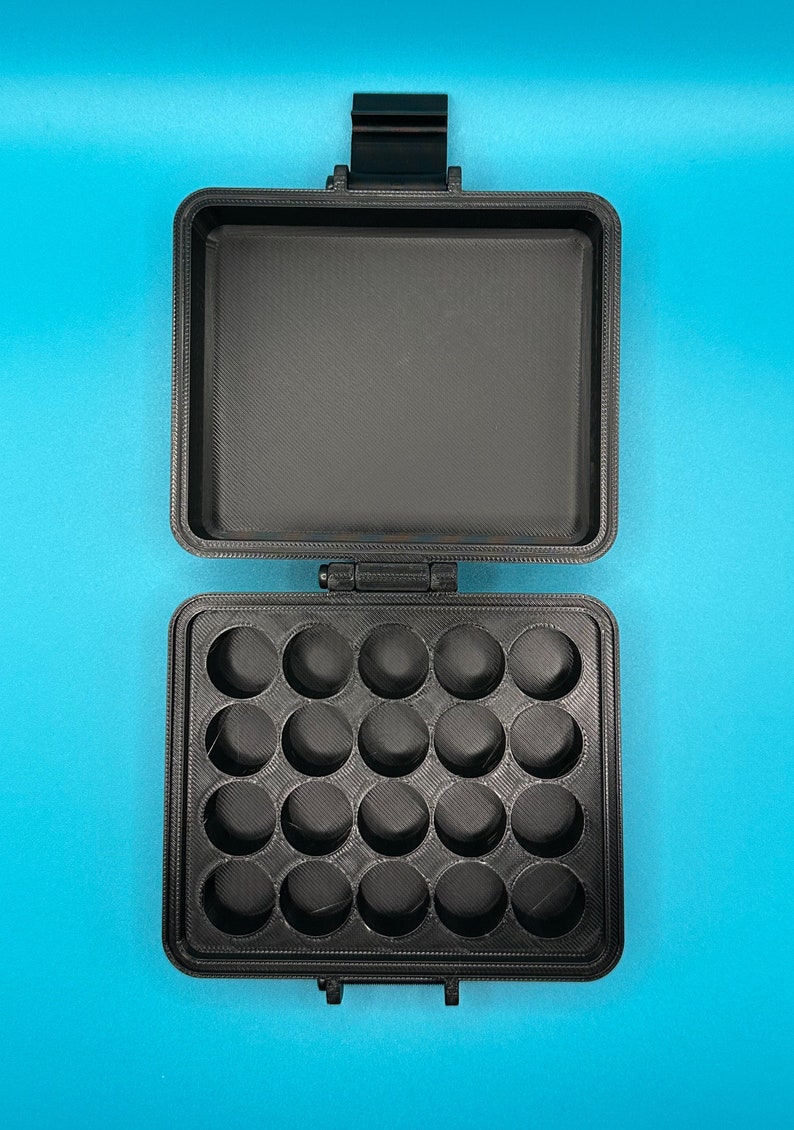 May include: A black rectangular storage case with a hinged lid. The interior base features a grid of twenty circular indentations arranged in four rows of five, while the interior lid is flat and smooth. The case has a textured matte finish.