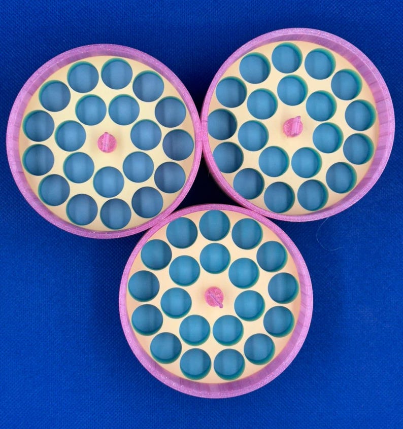 May include: Three circular storage organizers with pink outer rims and light yellow interior trays featuring multiple round holes. Each tray has a small pink center knob and reveals a blue base layer through the circular openings.