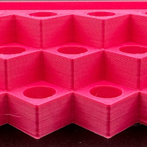 May include: A bright pink tiered display stand featuring multiple square-shaped compartments with circular openings. The organizer is constructed with a stepped design, providing three distinct levels for holding small items securely.