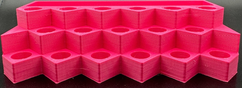 May include: A bright pink tiered display stand featuring multiple square-shaped compartments with circular openings. The organizer is constructed with a stepped design, providing three distinct levels for holding small items securely.