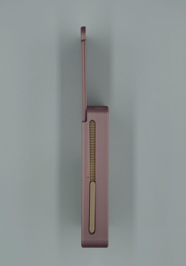 May include: A mauve-colored, rectangular lighter with a hinged lid. The lighter has a vertical, ribbed design on the side and a sliding mechanism. The lid is open, revealing the interior components. The lighter is set against a light blue background.