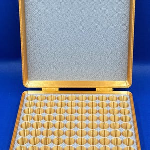 K&ouml;nnte beinhalten: Ein goldfarbener Kunststoff-Aufbewahrungskasten mit einem Scharnierdeckel und einem Schaumstoff-Innenraum mit mehreren runden F&auml;chern. Der Kasten ist ge&ouml;ffnet und der Schaumstoff-Innenraum ist sichtbar.