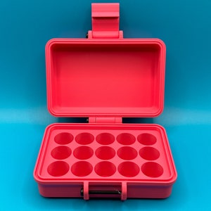 Puede incluir: Una caja de plástico de color rosa brillante con una tapa con bisagras y un asa. La caja tiene 12 compartimentos redondos.