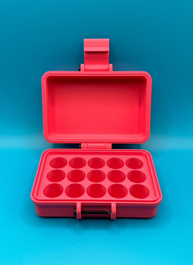 Puede incluir: Un estuche de almacenamiento rectangular de color rosa brillante con la tapa abierta y una base que presenta quince compartimentos circulares dispuestos en tres filas de cinco. El estuche incluye un mecanismo de cierre con bisagras y un peque&ntilde;o pasador de metal negro visible en la parte frontal.