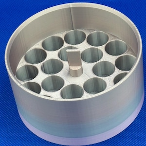 May include: A cylindrical, 3D-printed organizer with a gradient color finish transitioning from light purple to silver. The interior features a grid of nineteen circular holes and a central vertical tab for handling.