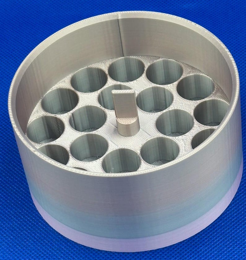 May include: A cylindrical, 3D-printed organizer with a gradient color finish transitioning from light purple to silver. The interior features a grid of nineteen circular holes and a central vertical tab for handling.