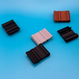 May include: A collection of five small, rectangular plastic clips featuring a wavy, corrugated surface texture. The set includes three black clips, one light gray clip, and one reddish-brown clip, all arranged in a scattered formation.