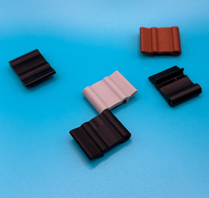 May include: A collection of five small, rectangular plastic clips featuring a wavy, corrugated surface texture. The set includes three black clips, one light gray clip, and one reddish-brown clip, all arranged in a scattered formation.