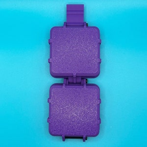 May include: A purple plastic sandwich press featuring two square, textured cooking plates connected by a central hinge mechanism with a vertical handle tab extending from the top, designed for creating sealed, toasted square sandwiches.