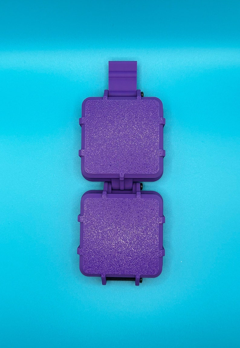 May include: A purple plastic sandwich press featuring two square, textured cooking plates connected by a central hinge mechanism with a vertical handle tab extending from the top, designed for creating sealed, toasted square sandwiches.