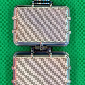 May include: A rectangular, two-part hinged case with a metallic, iridescent finish featuring a diagonal textured pattern. The object has a small, striped tab extending from the top edge and a secure latch mechanism connecting the two halves.