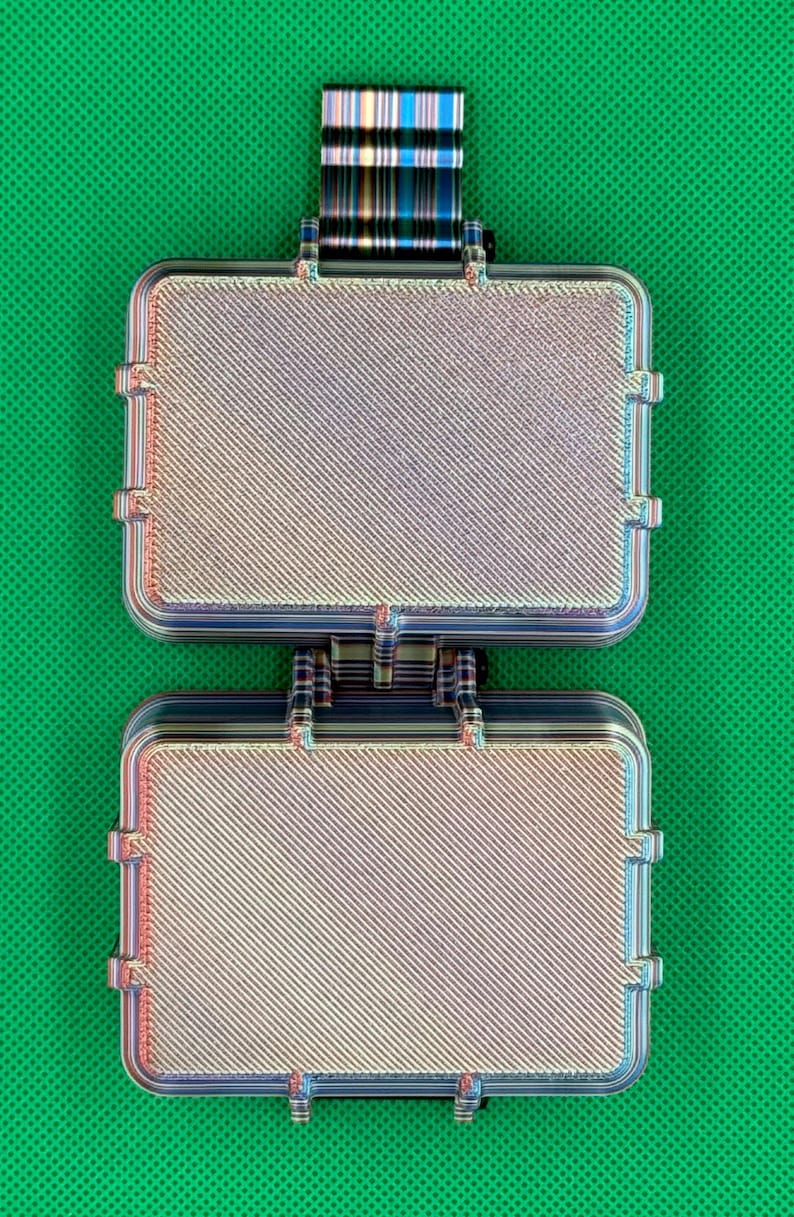 May include: A rectangular, two-part hinged case with a metallic, iridescent finish featuring a diagonal textured pattern. The object has a small, striped tab extending from the top edge and a secure latch mechanism connecting the two halves.