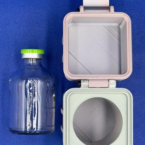May include: A clear glass vial with a bright green cap sits beside a two-part, 3D-printed square container. The top section is light pink with a square interior, and the bottom section is light green with a circular interior cutout.