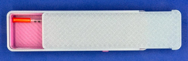 May include: A rectangular sliding case with a textured silver exterior and a bright pink interior. The case is partially open, revealing a small, clear plastic syringe with a vibrant orange cap resting inside the pink compartment.