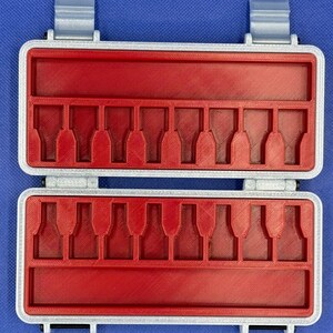 May include: A rectangular storage case with a silver-colored frame and a bright red interior insert. The insert features two rows of eight molded slots designed to hold small items, with a long horizontal compartment above and below each row.