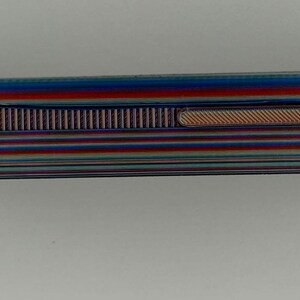 May include: A rectangular lighter with a striped pattern in shades of blue, red, and green. The lighter has a textured, ridged section and a small, oval-shaped indentation. The overall design is sleek and modern.