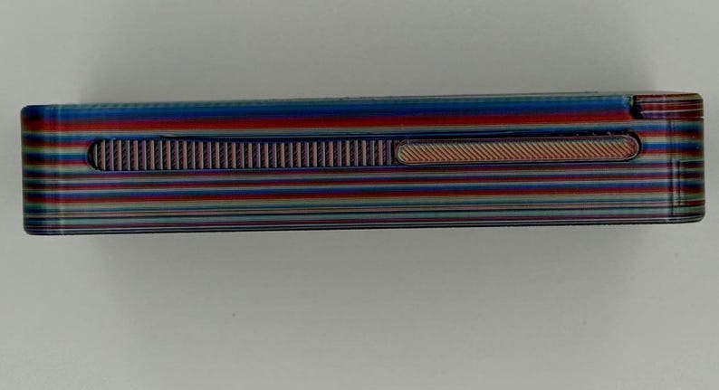 May include: A rectangular lighter with a striped pattern in shades of blue, red, and green. The lighter has a textured, ridged section and a small, oval-shaped indentation. The overall design is sleek and modern.
