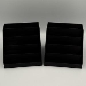 May include: Two black, tiered display stands. Each stand has a rectangular base and multiple horizontal shelves, creating a stepped appearance. The stands are made of a matte black material and are designed to hold and showcase items.