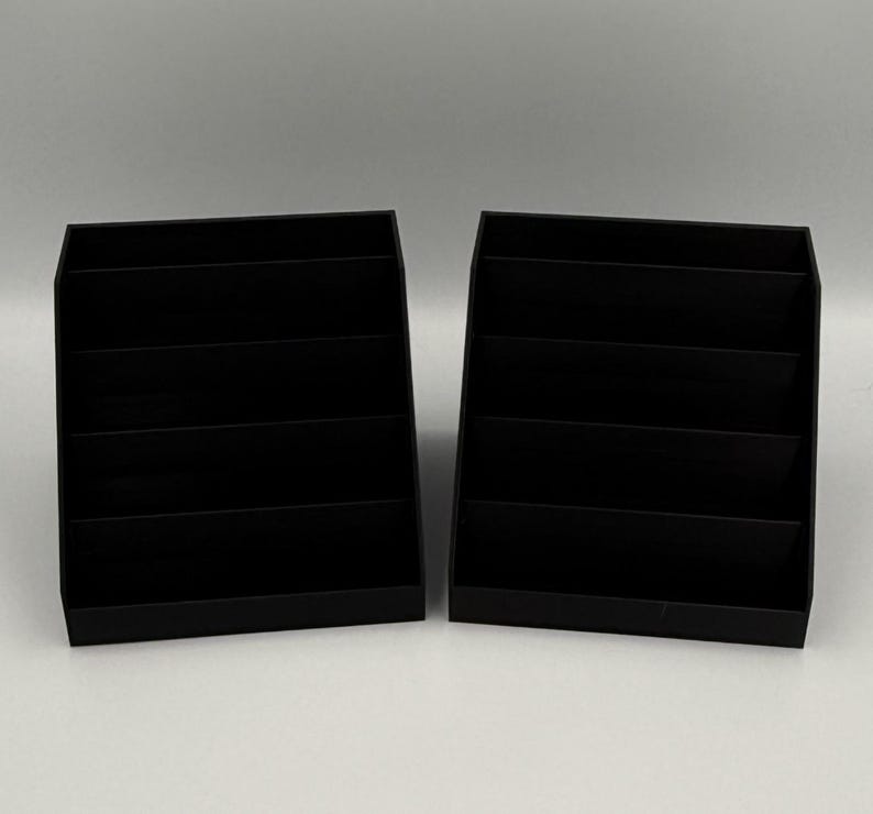 May include: Two black, tiered display stands. Each stand has a rectangular base and multiple horizontal shelves, creating a stepped appearance. The stands are made of a matte black material and are designed to hold and showcase items.