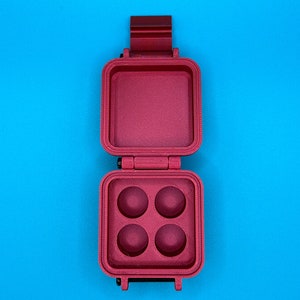 Puede incluir: Estuche cuadrado burdeos abierto con cuatro hendiduras circulares. El estuche está abierto, revelando el interior. El estuche está sobre un fondo azul brillante. El estuche tiene una tapa con bisagras y un pestillo.
