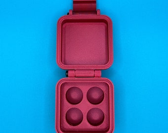 May include: A magenta, square-shaped, hinged container with a textured finish. The top lid is open, revealing a flat interior surface. The bottom section features four circular, concave indentations arranged in a two-by-two grid pattern.