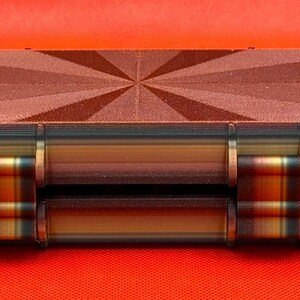 May include: A rectangular storage case featuring a dark brown lid with a textured sunburst pattern. The sides of the case display horizontal layers of brown, tan, and black, with two prominent hinges connecting the lid to the base.