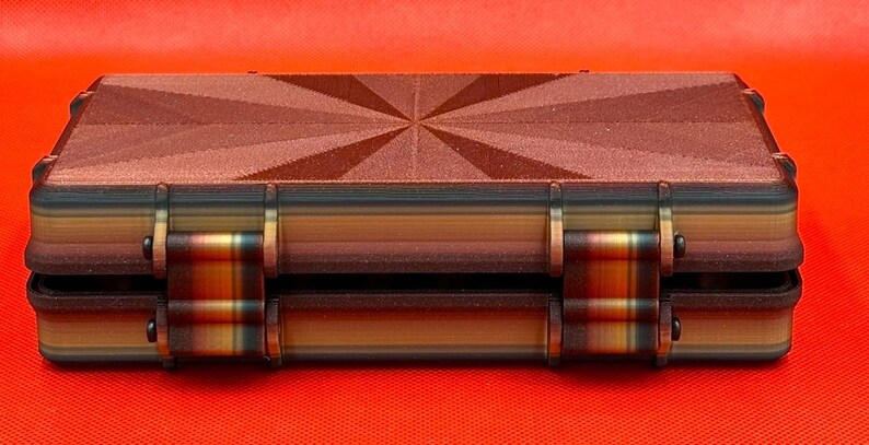 May include: A rectangular storage case featuring a dark brown lid with a textured sunburst pattern. The sides of the case display horizontal layers of brown, tan, and black, with two prominent hinges connecting the lid to the base.