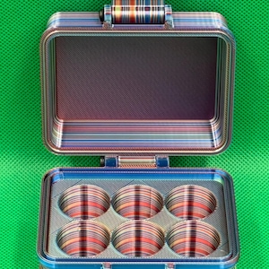 Puede incluir: Una pequeña caja de almacenamiento rectangular multicolor con seis compartimentos redondos. La caja está hecha de un material en capas con un patrón a rayas. La tapa está abierta, revelando los compartimentos vacíos.