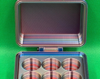 May include: A rectangular storage case with a hinged lid, featuring a multi-colored horizontal striped pattern. The interior base contains six circular compartments arranged in two rows of three. A black latch secures the front of the case.