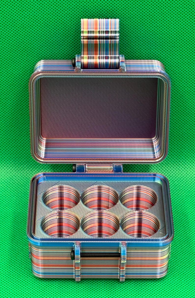 May include: A rectangular storage case with a hinged lid, featuring a multi-colored horizontal striped pattern. The interior base contains six circular compartments arranged in two rows of three. A black latch secures the front of the case.