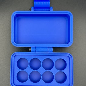 May include: A bright blue rectangular storage case with a hinged lid. The interior features a bottom section with eight circular cutouts arranged in two rows of four, and a top section that is open and hollow for storage.
