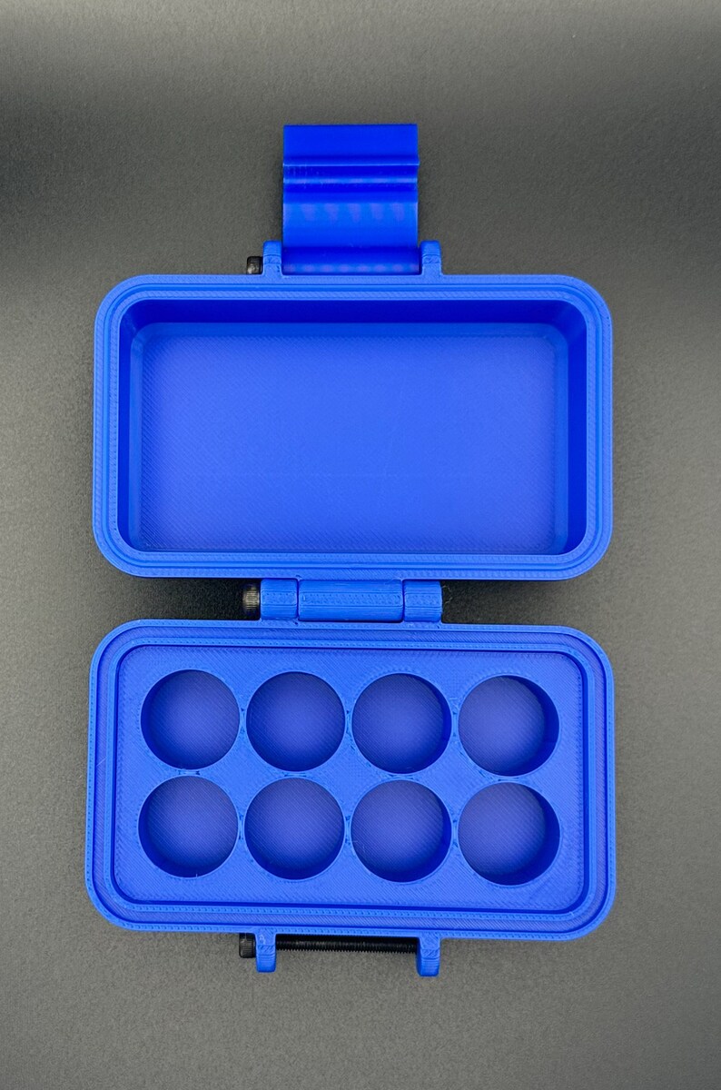 May include: A bright blue rectangular storage case with a hinged lid. The interior features a bottom section with eight circular cutouts arranged in two rows of four, and a top section that is open and hollow for storage.