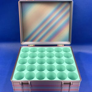 May include: A rectangular storage case with a multi-colored, iridescent striped exterior and a hinged lid. The interior features a light mint green insert with thirty-five circular cutouts arranged in a grid for organizing small items.