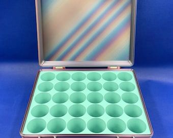 May include: A rectangular storage case with a multi-colored, iridescent striped exterior and a hinged lid. The interior features a light mint green insert with thirty-five circular cutouts arranged in a grid for organizing small items.