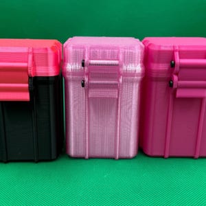 Puede incluir: Tres cajas de almacenamiento rectangulares con tapas abatibles y cierres de pestillo. La primera es negra con tapa y pestillo rosa brillante. La segunda es rosa claro transl&uacute;cido. La tercera es rosa brillante. Todas presentan estr&iacute;as verticales.