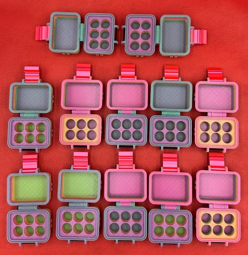 May include: A collection of rectangular, hinged storage cases arranged in rows. Each case features a bright pink latch, a multi-colored frame, and an interior divided into a solid compartment and a tray with six circular indentations.