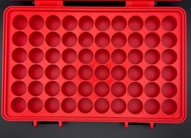 K&ouml;nnte beinhalten: Ein leuchtend roter, rechteckiger Aufbewahrungskoffer mit einem Raster aus sechsundf&uuml;nfzig kreisf&ouml;rmigen, vertieften F&auml;chern. Der Koffer verf&uuml;gt &uuml;ber einen Klappdeckel mit zwei schwarzen Verschl&uuml;ssen an der Unterkante.