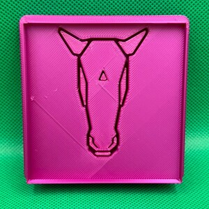 K&ouml;nnte beinhalten: Ein rosa quadratischer 3D-Druck mit einem Pferdekopf-Design in der Mitte.