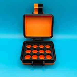 May include: An orange and black plastic storage case with a hinged lid and 12 round compartments. The case is open and empty.