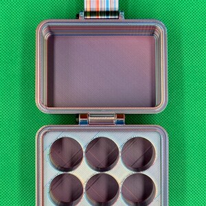 May include: A rectangular, multi-colored, 3D-printed storage case with a hinged lid. The interior features a flat compartment on the top half and six circular recessed slots on the bottom half. The case displays a layered, iridescent finish.