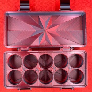 Puede incluir: Un estuche de almacenamiento rectangular de color rojo oscuro impreso en 3D con tapa abatible. El interior de la tapa presenta un patr&oacute;n de estrella geom&eacute;trico, mientras que la base contiene diez compartimentos circulares dispuestos en dos filas de cinco, todos con un acabado texturizado.