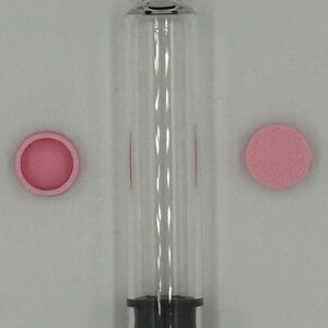 May include: A clear glass cylindrical vial with a pink cap attached to the top, featuring a black rubber stopper at the base. Two additional pink circular caps are positioned on either side of the vial, one showing the interior and one showing the exterior.