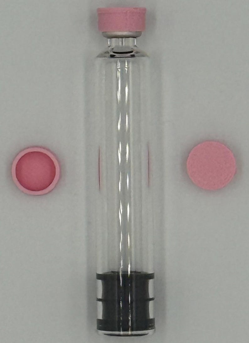 May include: A clear glass cylindrical vial with a pink cap attached to the top, featuring a black rubber stopper at the base. Two additional pink circular caps are positioned on either side of the vial, one showing the interior and one showing the exterior.
