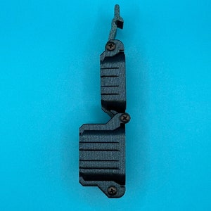 K&ouml;nnte beinhalten: Ein dunkelgraues, 3D-gedrucktes Klappgeh&auml;use mit einer texturierten, gerippten Au&szlig;enfl&auml;che. Das Objekt hat eine vertikale Ausrichtung mit einem oberen Verschlussmechanismus und drei kleinen, dunklen Metallschrauben.
